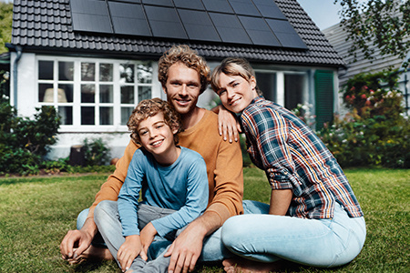 Ein Vater hebt seinen Sohn in die Luft, während der Sohn einen Fußball hält. Beide lachen und stehen im Garten vor einem Haus mit Solarpanels auf dem Dach. Der Garten ist grün, und der Himmel ist klar und blau, was eine fröhliche, sonnige Atmosphäre vermi